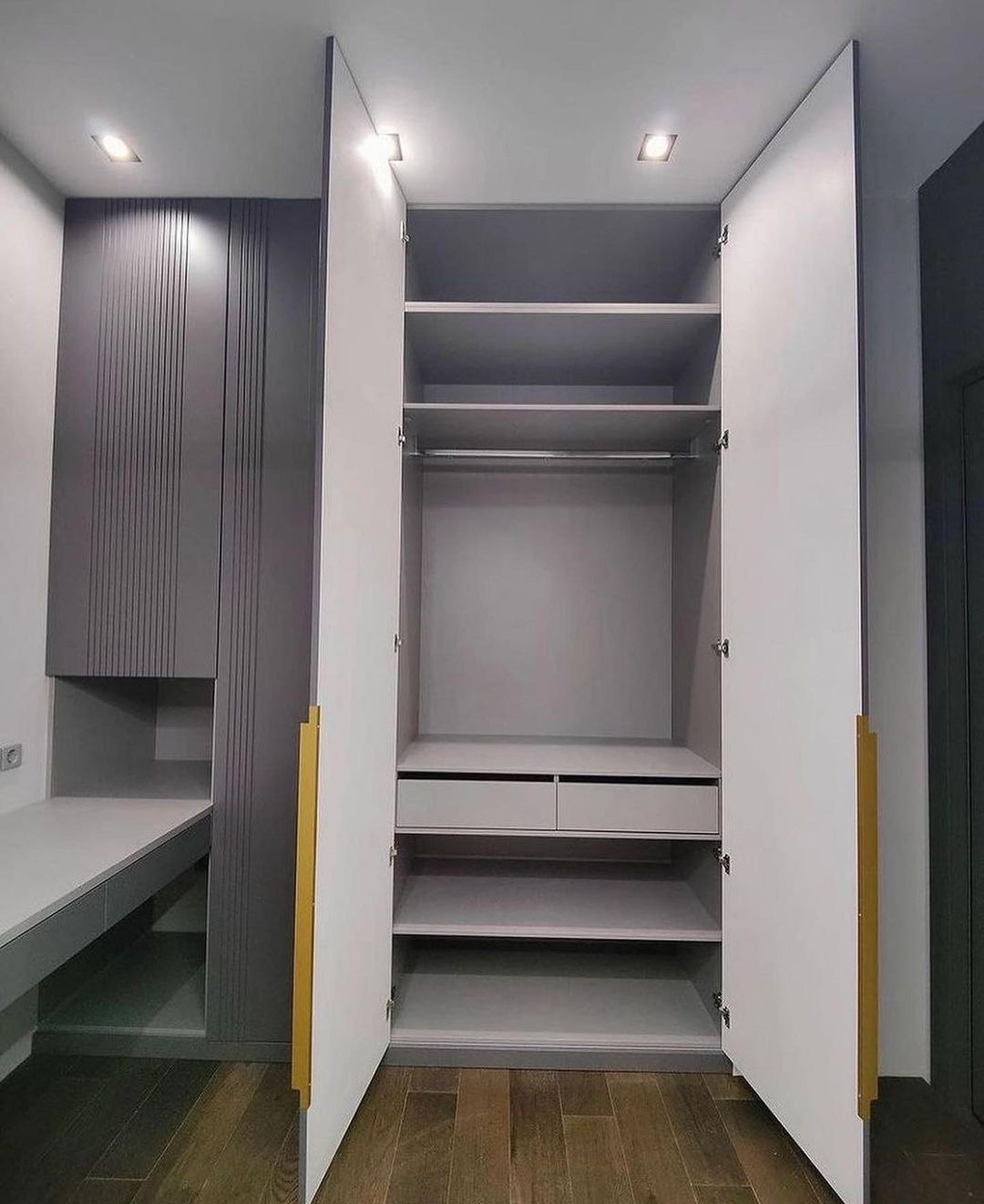 Szafa wnękowa z białymi drzwiami i złotymi uchwytami, widoczne półki i szuflady, obok biurko z szarymi frontami i pionowymi żłobieniami, oświetlenie punktowe sufitowe.