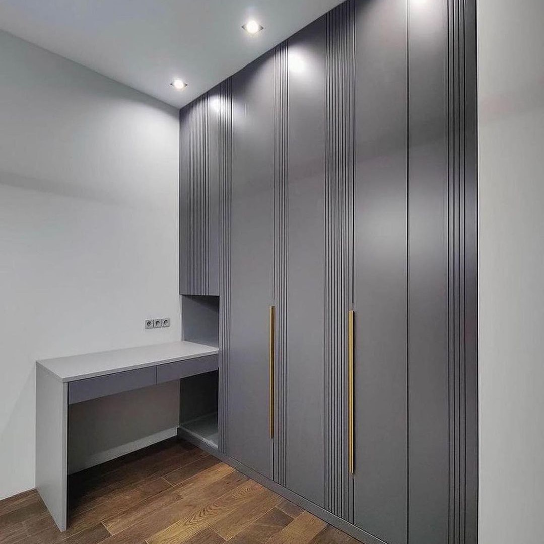 Szara szafa wnękowa z pionowymi frezowaniami i złotymi uchwytami, zintegrowana z biurkiem o minimalistycznym designie, na drewnianej podłodze.