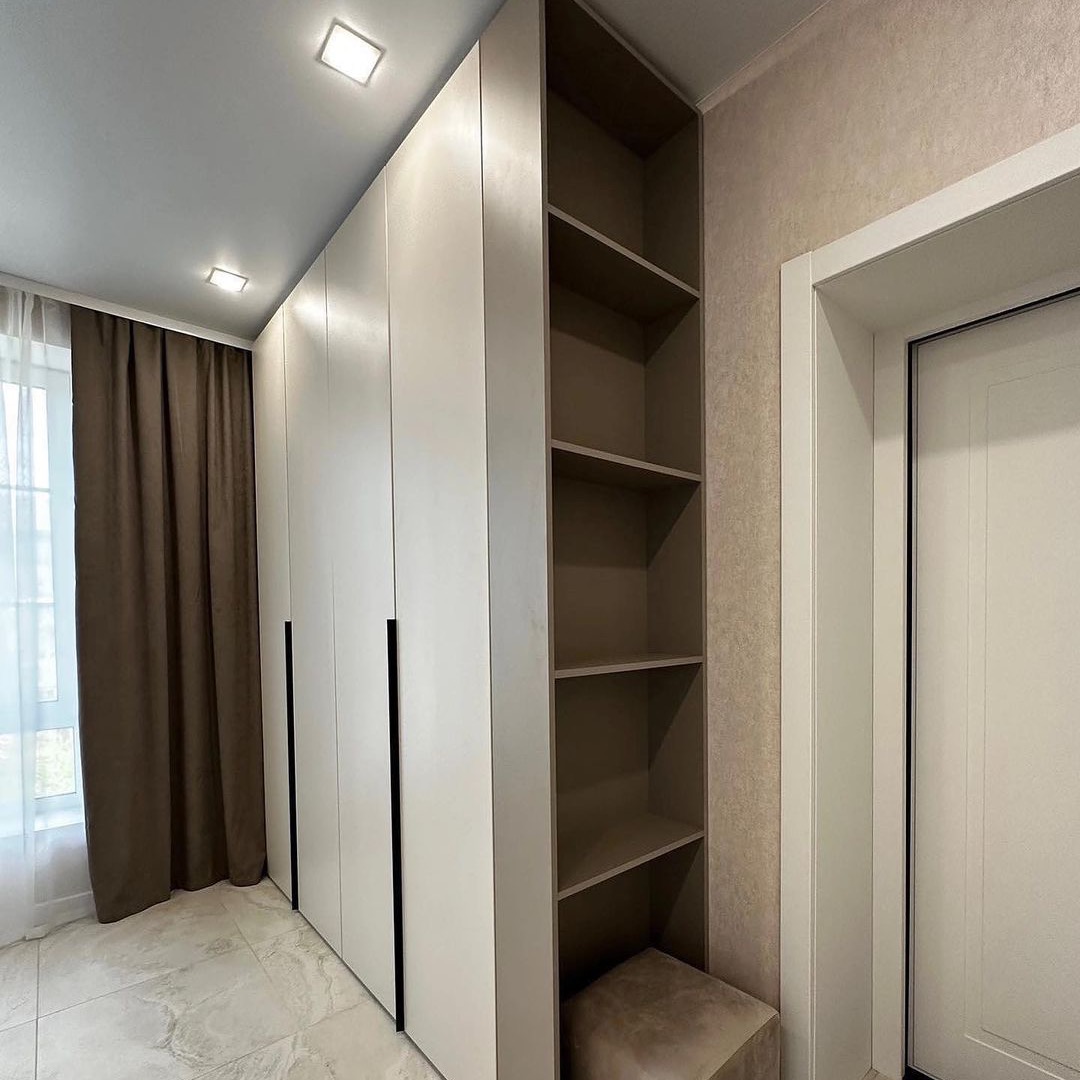 Biała szafa wnękowa z czarnymi uchwytami, przylegająca do otwartych półek w kolorze taupe, uzupełniona pufem w tym samym odcieniu, na tle beżowej ściany i okna z brązowymi zasłonami.