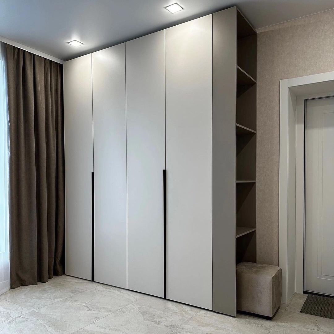 Duża, jasna szafa wnękowa z przesuwnymi drzwiami i czarnymi uchwytami, uzupełniona otwartymi półkami i pufą w neutralnych barwach.