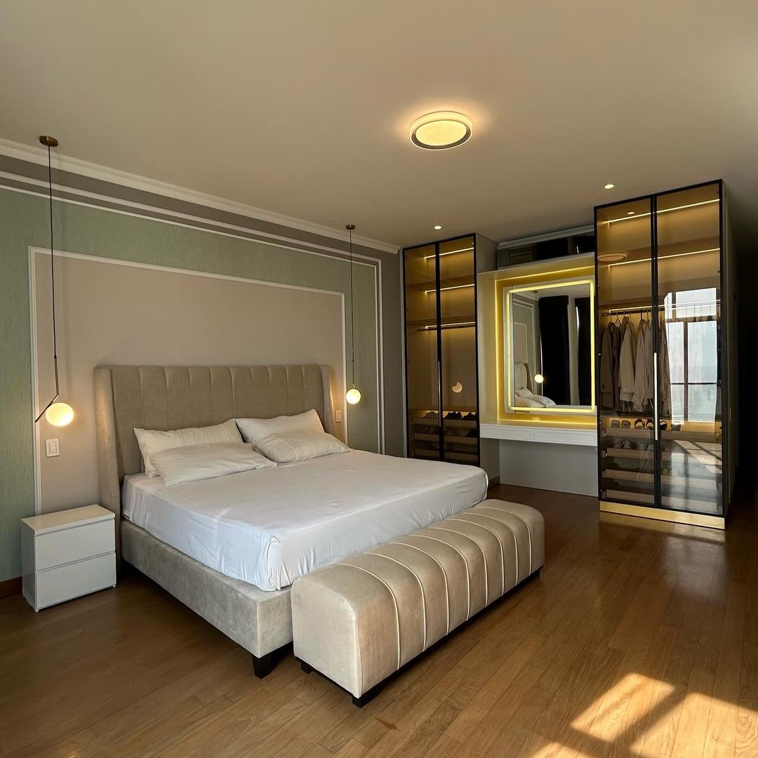 Nowoczesna sypialnia z łóżkiem z tapicerowanym wezgłowiem, beżową ławeczką, szafką nocną i podświetlaną garderobą z przeszklonymi drzwiami oraz toaletką z lustrem.