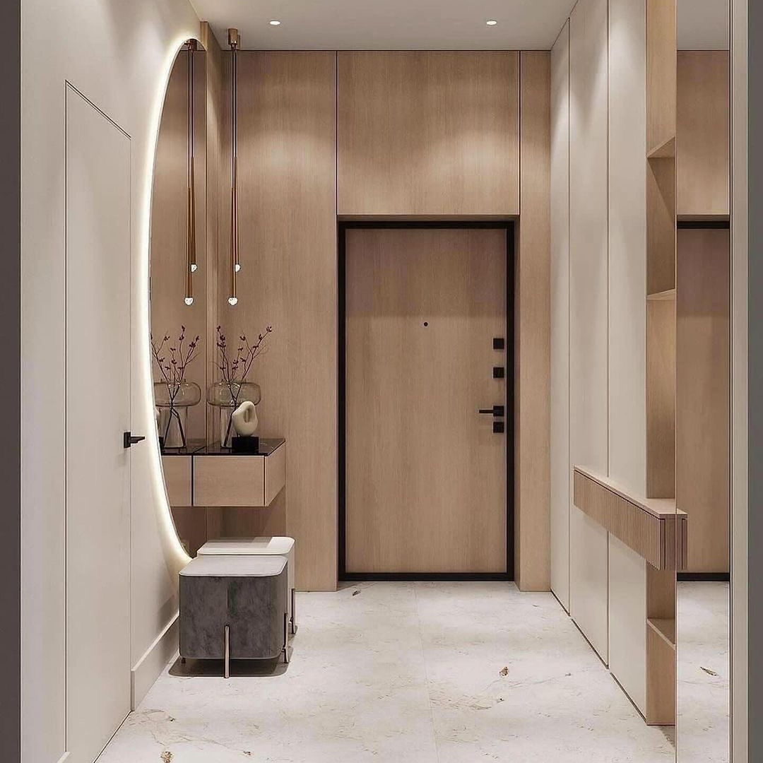 Nowoczesny przedpokój z podświetlanym owalnym lustrem, minimalistyczną konsolą z dekoracjami, tapicerowanym siedziskiem i zabudową meblową w odcieniach beżu i bieli.