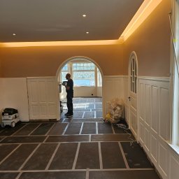 MARIUSZ BARZOWSKI DrewEfekt - Wnętrze w trakcie wykończenia: montaż paneli ściennych, zabezpieczona podłoga, widoczny pracownik z narzędziem i odkurzacz przemysłowy. Oświetlenie sufitowe.