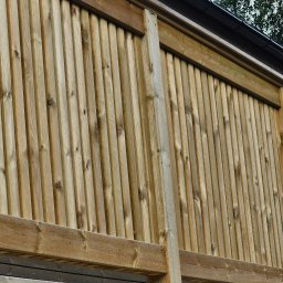 MARIUSZ BARZOWSKI DrewEfekt - Drewniana elewacja domu z pionowymi deskami, widok z dołu, fragment dachu z rynną w tle, naturalne oświetlenie, widoczne słoje drewna.