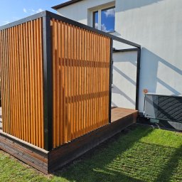 MARIUSZ BARZOWSKI DrewEfekt - Drewniana pergola z pionowymi lamelami w kolorze jasnego drewna, z czarną ramą, przylegająca do białej ściany budynku. Widoczny fragment tarasu i trawnik.