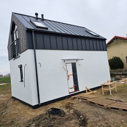 MARIUSZ BARZOWSKI DrewEfekt - Nowoczesny dom modułowy z białą elewacją i czarnym dachem, widok z zewnątrz, teren wokół w trakcie prac wykończeniowych, palety i wykop w ziemi.