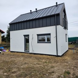 MARIUSZ BARZOWSKI DrewEfekt - Nowoczesny, dwukondygnacyjny dom z białą elewacją i ciemnym, metalowym dachem. Widoczne okna z czarnymi ramami i elementy wykończeniowe. Teren wokół domu częściowo zagospodarowany.