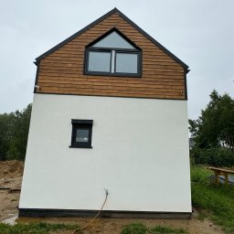 MARIUSZ BARZOWSKI DrewEfekt - Nowoczesny dom z białą elewacją i drewnianym wykończeniem poddasza, okno trójkątne. Widok z boku, na pierwszym planie ziemia i trawa. Minimalistyczny design.