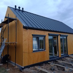 MARIUSZ BARZOWSKI DrewEfekt - Nowoczesny dom szkieletowy z drewnianą elewacją i czarnym dachem z blachy na etapie wykończenia. Widoczne rusztowania i pracownik przy elewacji.