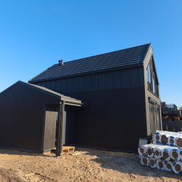 MARIUSZ BARZOWSKI DrewEfekt - Nowoczesny, ciemny dom z drewnianą elewacją i ciemną dachówką. Na pierwszym planie ocieplenie w rolkach. Widok z zewnątrz, w słoneczny dzień.