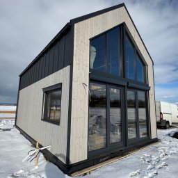 MARIUSZ BARZOWSKI DrewEfekt - Nowoczesny dom modułowy z dużymi oknami, wykończony drewnem i czarną blachą, na zasypanym śniegiem terenie. Widoczny biały bus i elementy konstrukcyjne.