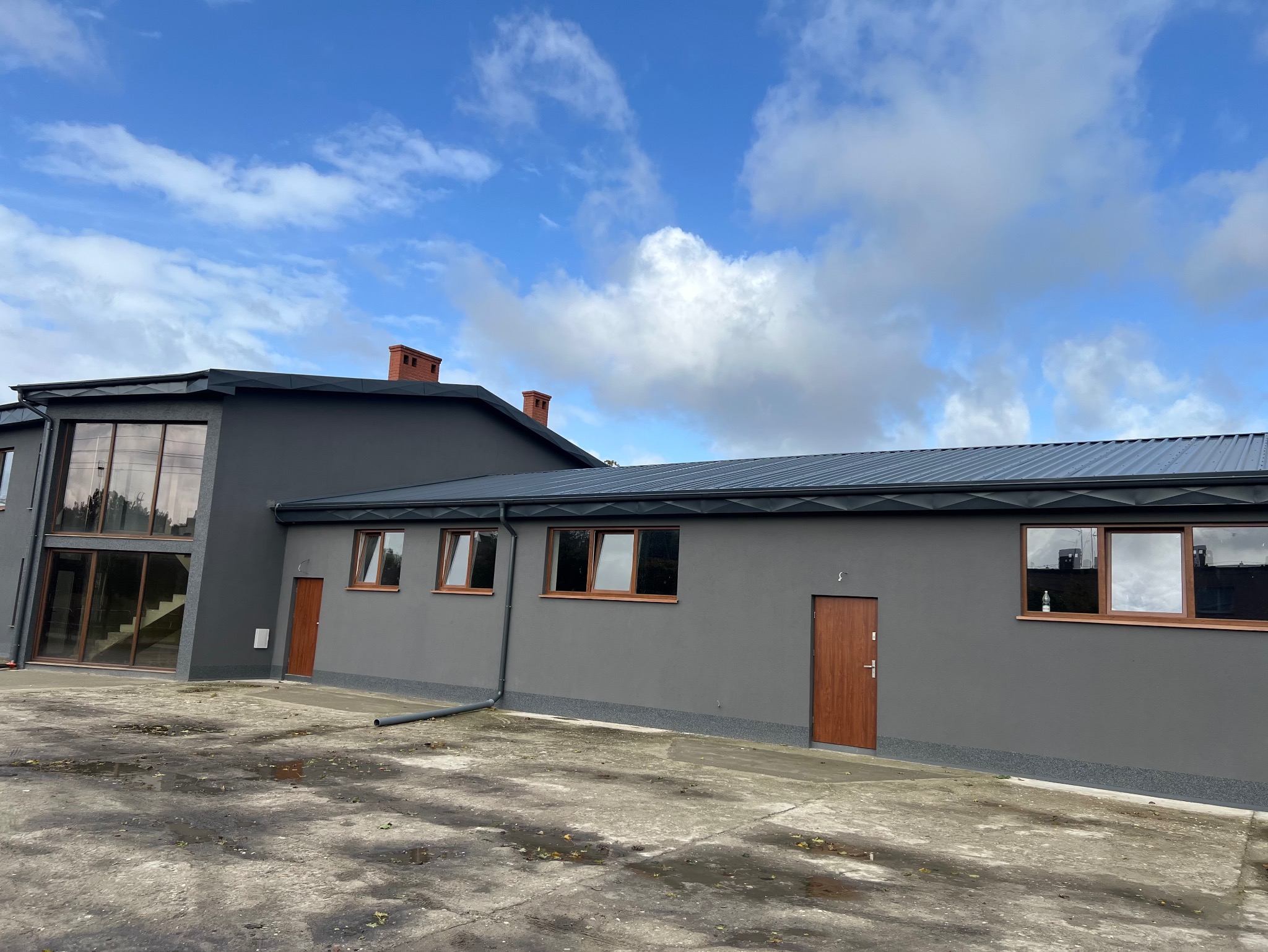 Szary budynek z ciemnym dachem z blachy trapezowej, brązowymi oknami i drzwiami. Widoczne kominy z cegły i fragment betonowego placu przed budynkiem.