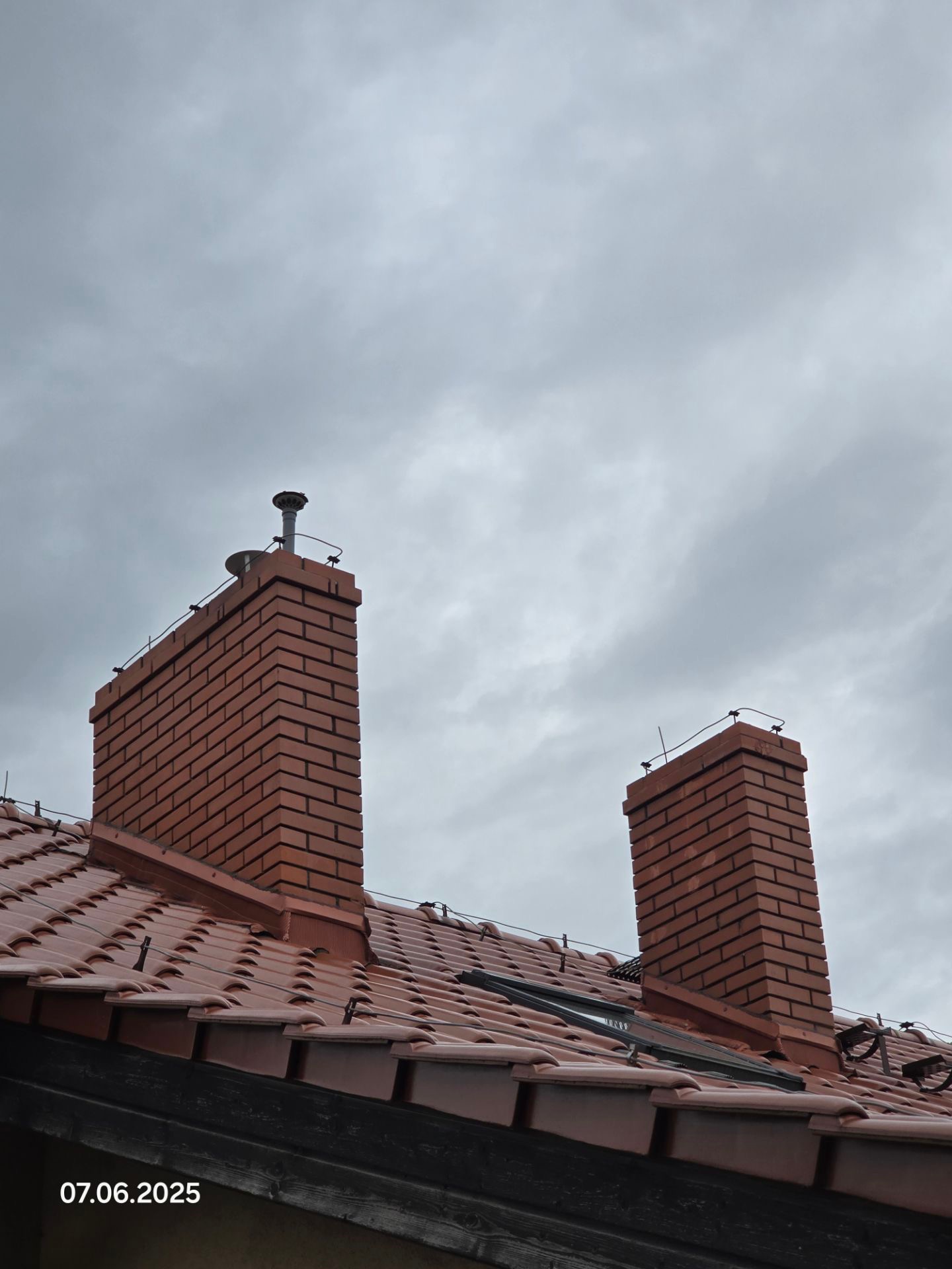 Czerwony dach z dwoma kominami z cegły pod pochmurnym niebem. Widoczne okno dachowe i elementy instalacji odgromowej. Data 07.06.2025 w lewym dolnym rogu.