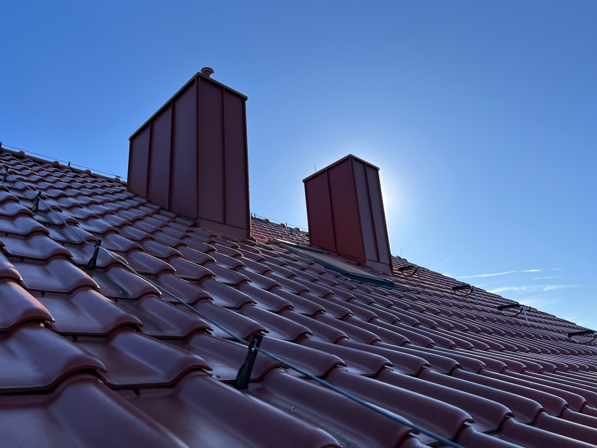 Czerwona dachówka ceramiczna i blaszane kominy na tle błękitnego nieba we Wrześni. Widok z dołu, podkreślający geometrię dachu i precyzję wykonania.