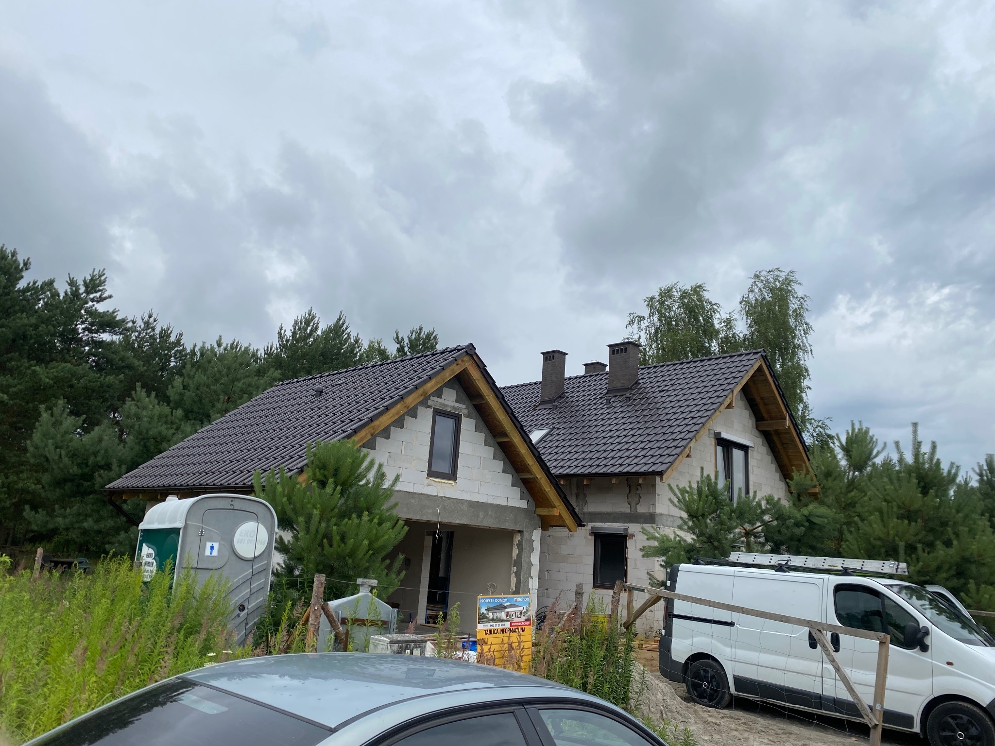 Dom w trakcie budowy z ciemnym dachem i widocznymi elementami konstrukcyjnymi. Na pierwszym planie fragment samochodu i przenośna toaleta.