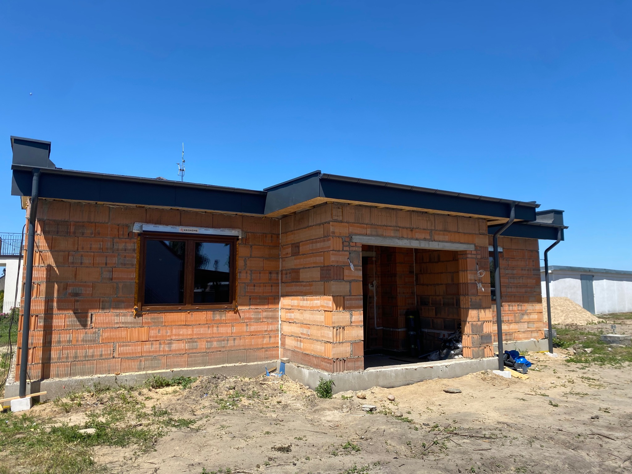 Surowy stan budynku z czerwonej cegły z zamontowaną stolarką okienną i elementami dachu w kolorze grafitowym, na tle błękitnego nieba.