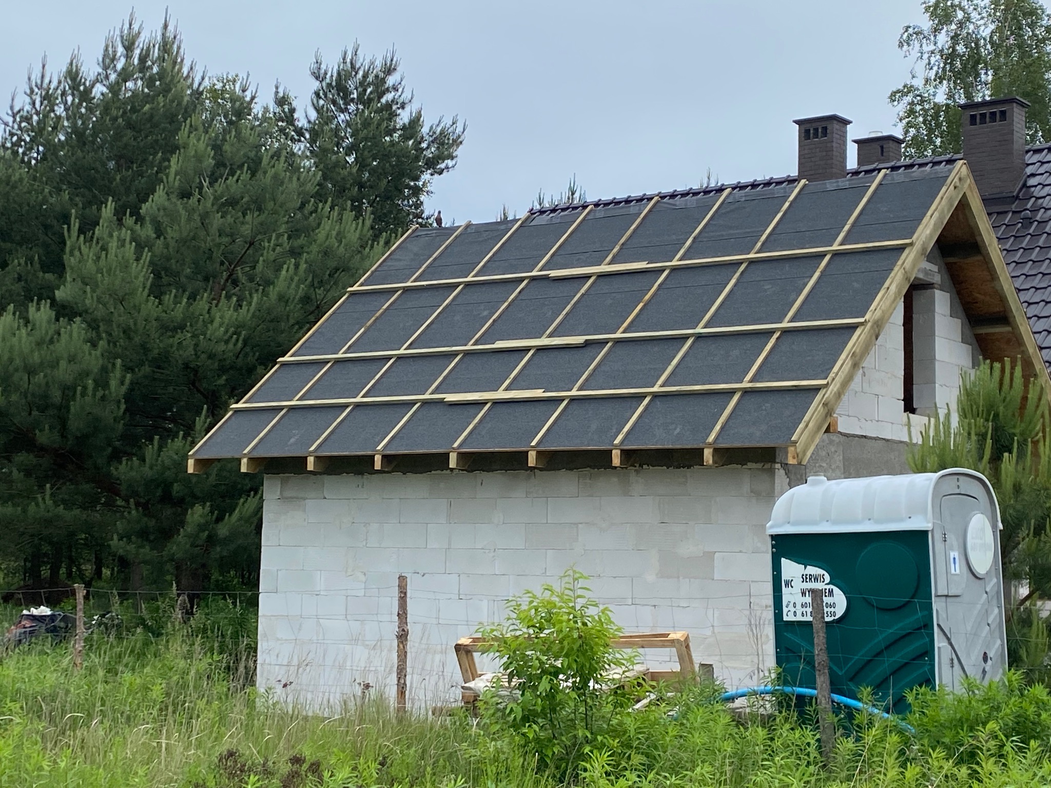 Budowa dachu: czarna papa na łatach na surowej konstrukcji domu z białego bloczka, widok z zewnątrz, zieleń w tle, przenośna toaleta obok budynku.