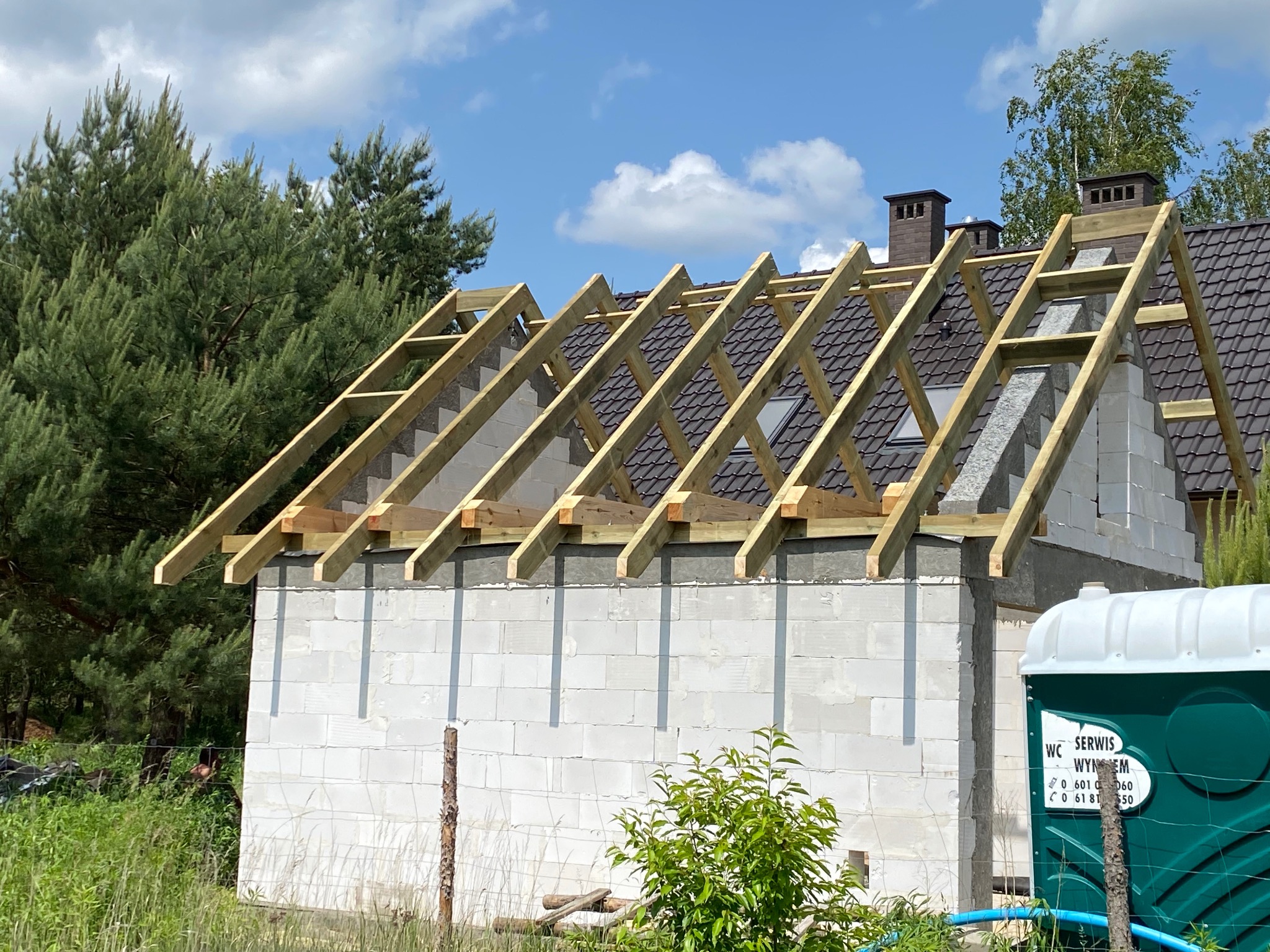 Budowa dachu z drewnianą konstrukcją na murowanym domu w słoneczny dzień. Widoczny fragment gotowego dachu z brązową dachówką i kominy. Na pierwszym planie toaleta przenośna.