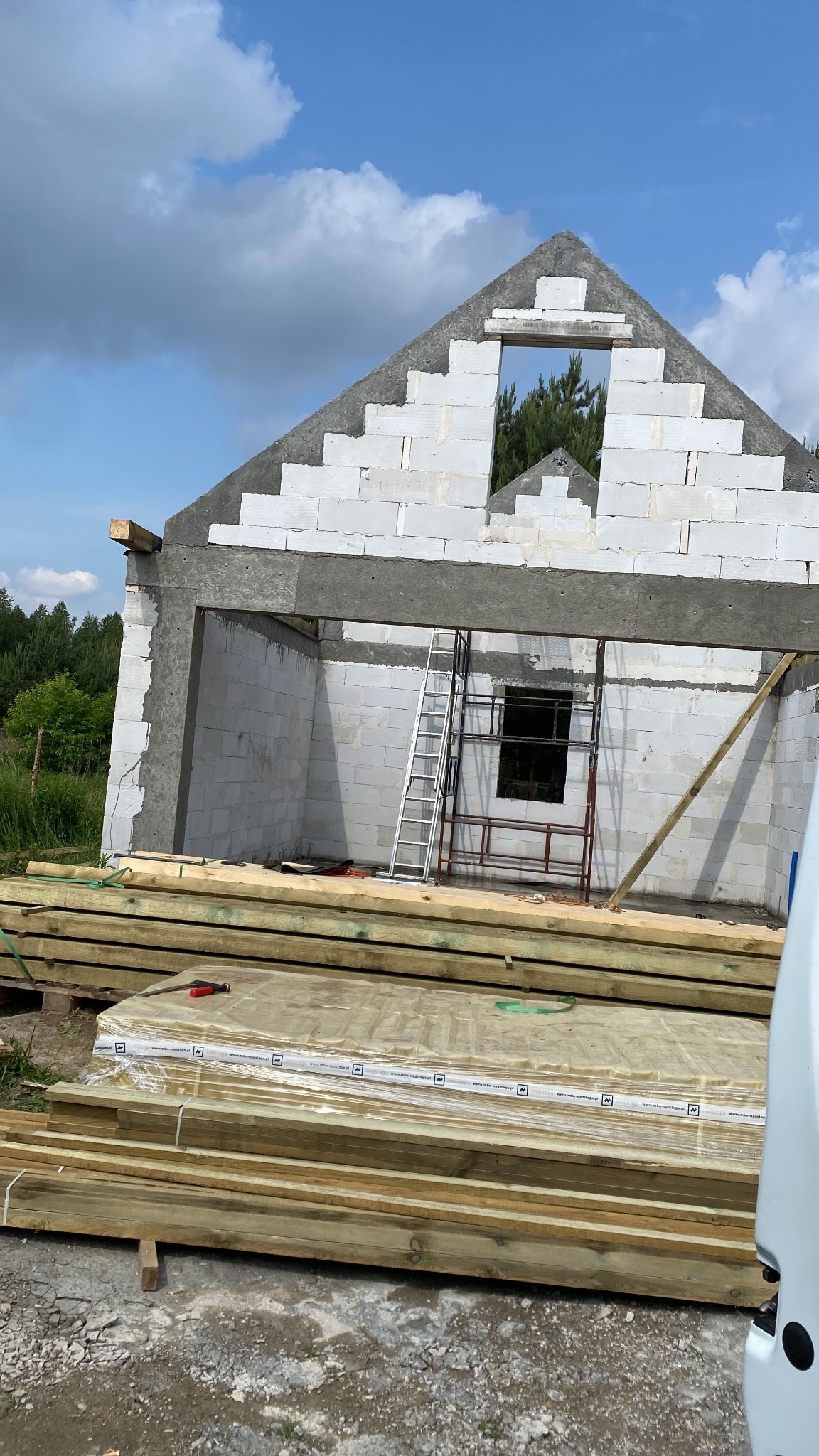 Budowa domu z białych bloczków, widoczne rusztowanie i drewniane elementy konstrukcyjne. W tle zieleń drzew i błękitne niebo z chmurami.