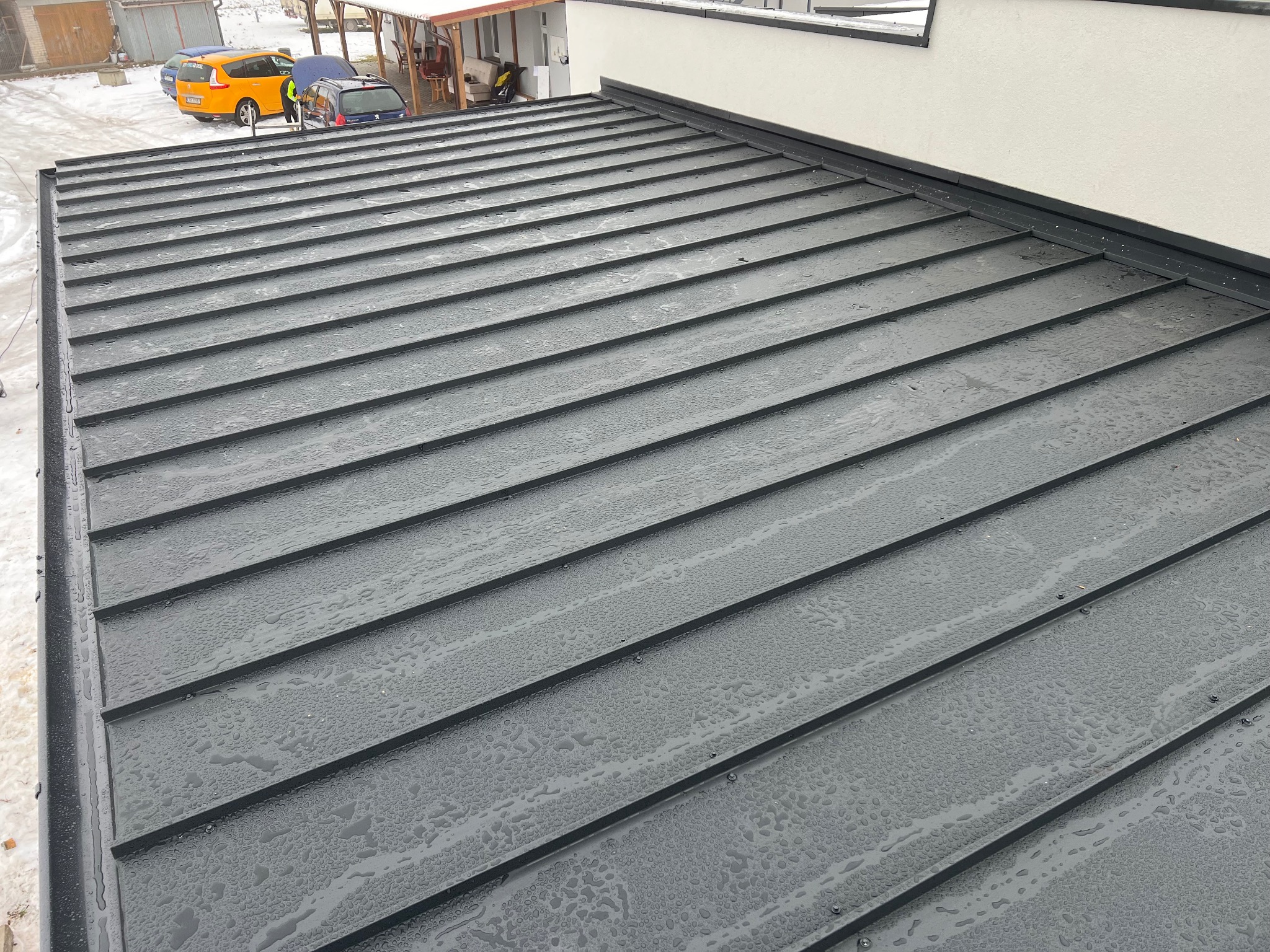 Nowoczesny, grafitowy dach z blachy na budynku jednorodzinnym, pokryty kroplami wody. W tle zaparkowane samochody i fragment ośnieżonego terenu.