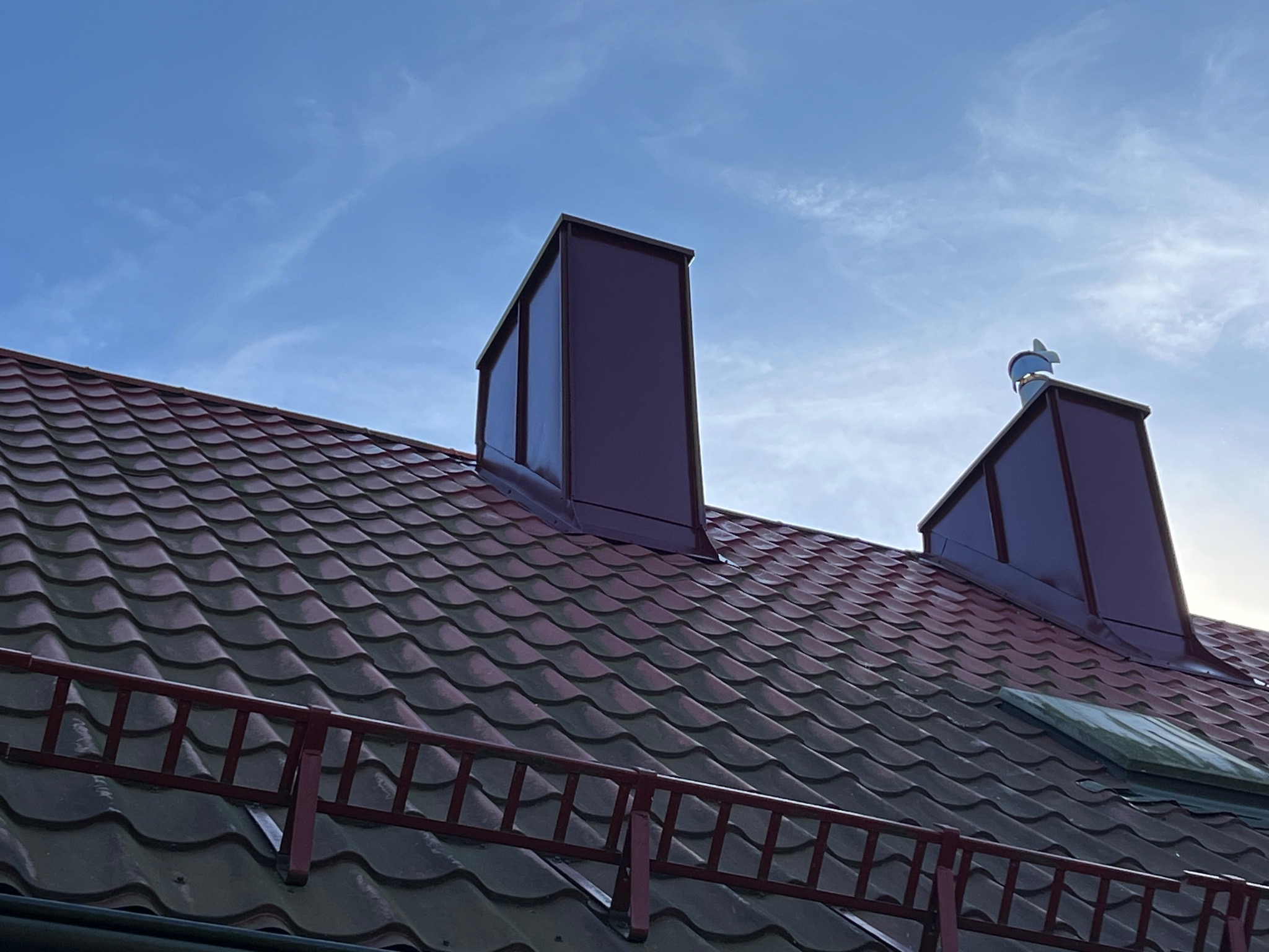 Czerwono-brązowy dach z blachodachówki z dwoma kominami i barierkami śniegowymi na tle błękitnego nieba z delikatnymi chmurami. Widoczny wiatrowskaz.