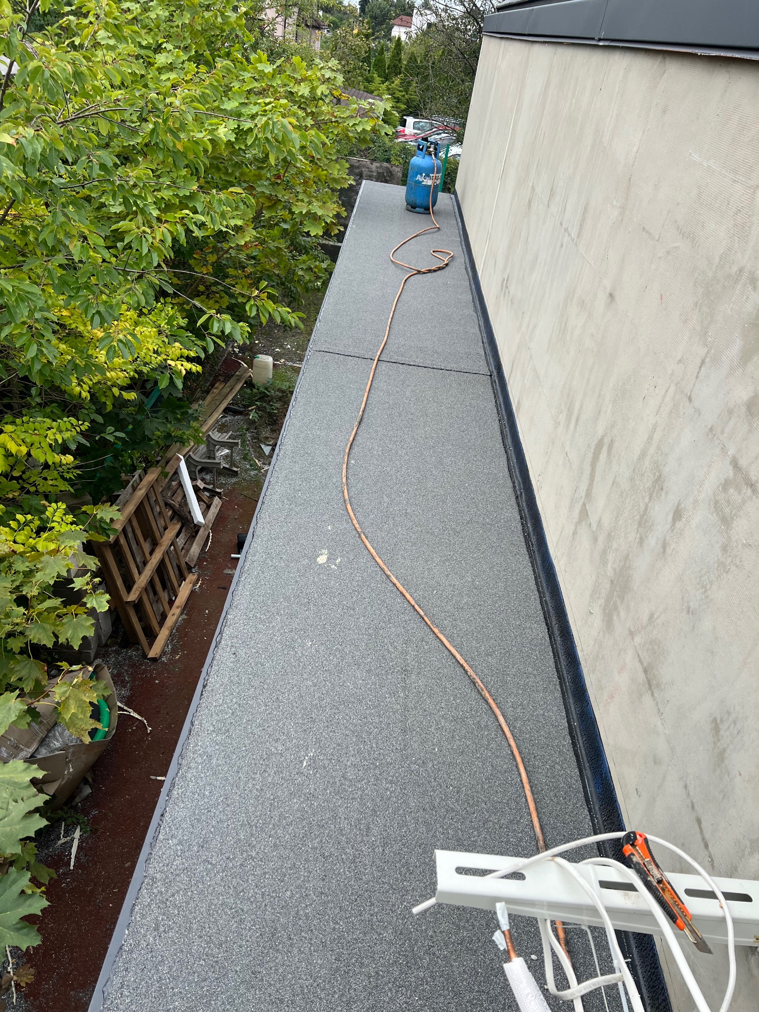 Szary dach pokryty papą termozgrzewalną, widoczny przewód gazowy i butla. Na pierwszym planie narzędzia dekarskie. Ujęcie z góry, z boku widoczna ściana budynku i zieleń.
