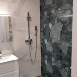 Nowoczesna łazienka z marmurowymi płytkami w odcieniach bieli i zieleni, chromowana armatura prysznicowa, biała szafka pod umywalką i lustro.