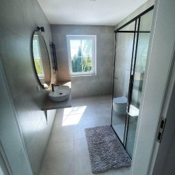 REMONT-LUX - Nowoczesna łazienka z szarymi płytkami, kabiną prysznicową typu walk-in z czarnymi ramami, owalnym lustrem, umywalką nablatową i grzejnikiem drabinkowym. Dywanik w odcieniach szarości leży...