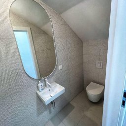 REMONT-LUX - Mała łazienka na poddaszu z umywalką, owalnym lustrem i wiszącą toaletą, wykończona szarą mozaiką na ścianach i szarą terakotą na podłodze. Na umywalce stoi butelka z mydłem.