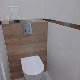 REMONT-LUX - Nowoczesna toaleta wisząca w łazience z białymi płytkami i drewnopodobną zabudową, chromowana listwa dekoracyjna, szara podłoga z płytek.