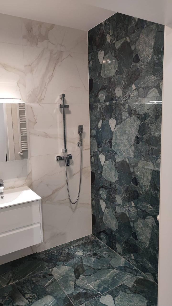 Nowoczesna łazienka z marmurowymi płytkami w odcieniach bieli i zieleni, chromowana armatura prysznicowa, biała szafka pod umywalką i lustro.