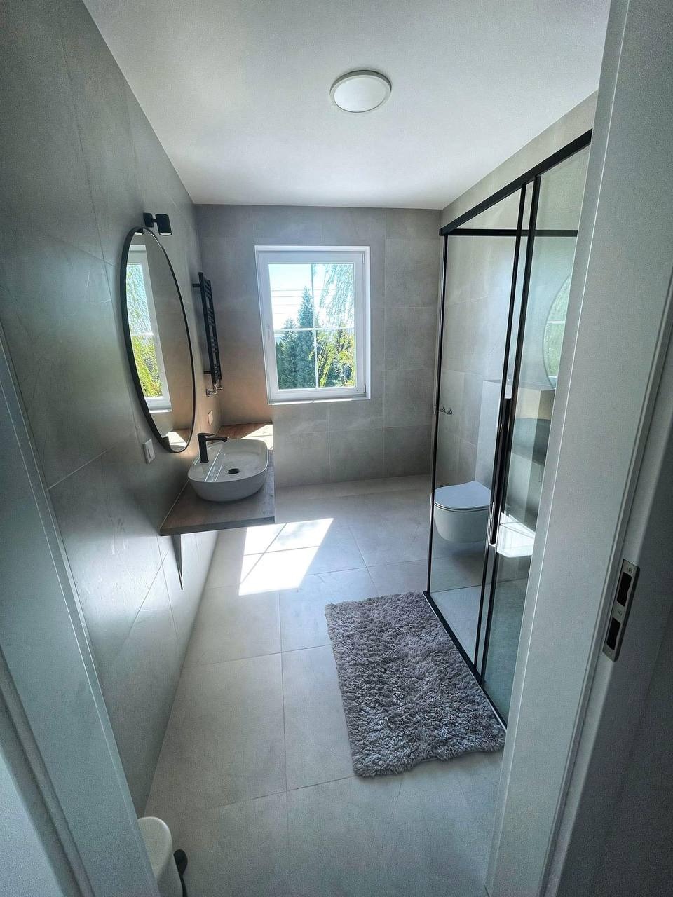 Nowoczesna łazienka z szarymi płytkami, kabiną prysznicową typu walk-in z czarnymi ramami, owalnym lustrem, umywalką nablatową i grzejnikiem drabinkowym. Dywanik w odcieniach szarości leży...