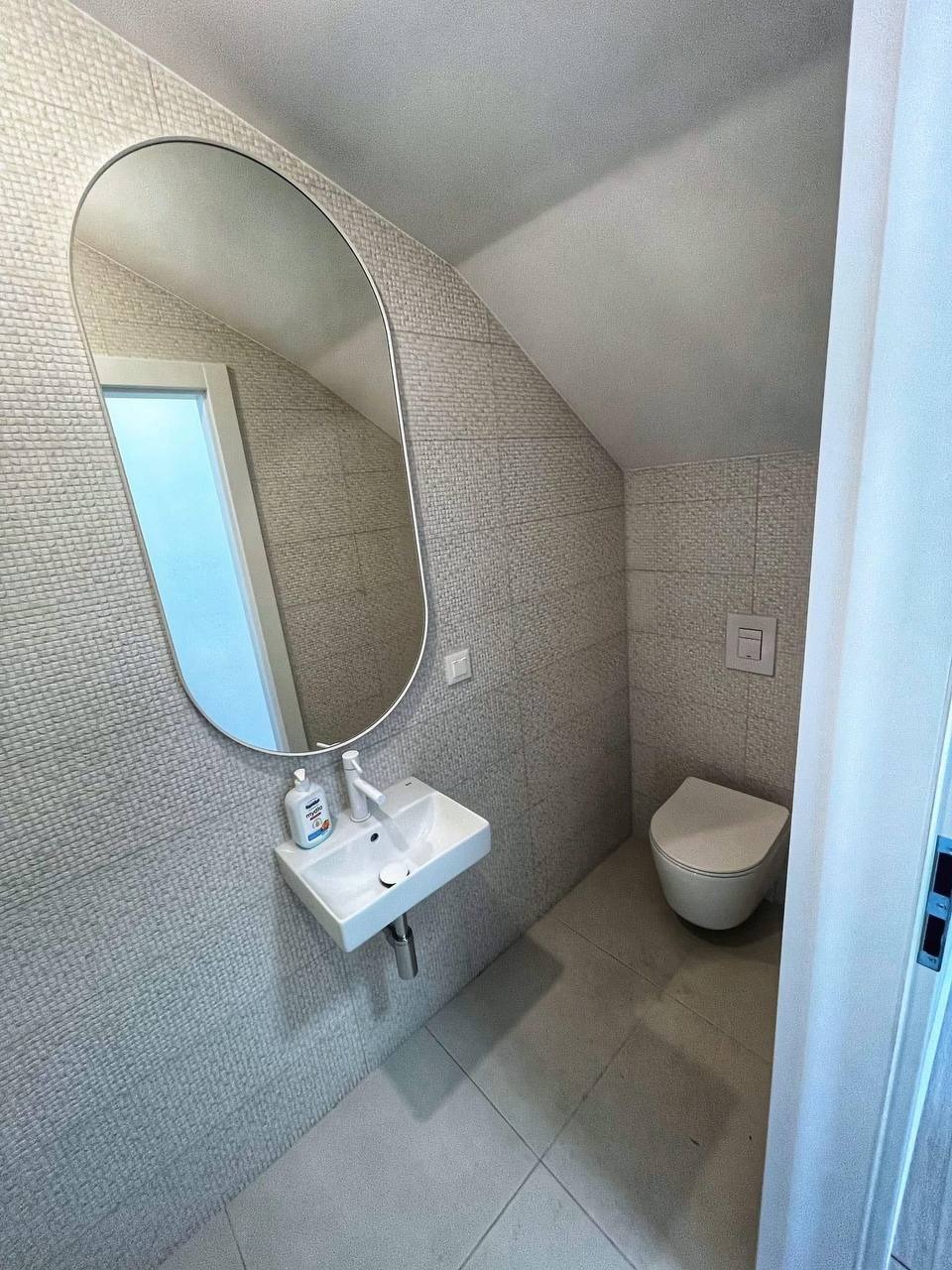 Mała łazienka na poddaszu z umywalką, owalnym lustrem i wiszącą toaletą, wykończona szarą mozaiką na ścianach i szarą terakotą na podłodze. Na umywalce stoi butelka z mydłem.