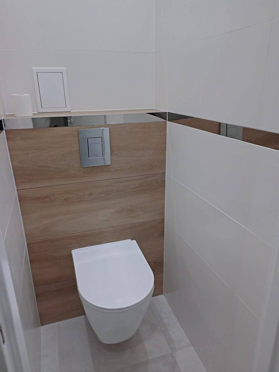Nowoczesna toaleta wisząca w łazience z białymi płytkami i drewnopodobną zabudową, chromowana listwa dekoracyjna, szara podłoga z płytek.