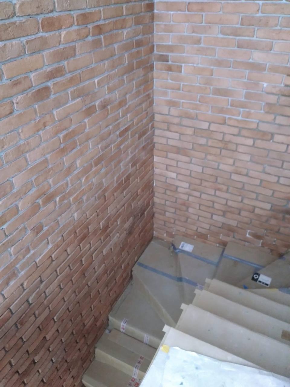Ściana wyłożona cegłą klinkierową w rogu pomieszczenia, z widocznymi schodami w kartonowych opakowaniach, przygotowanymi do montażu.