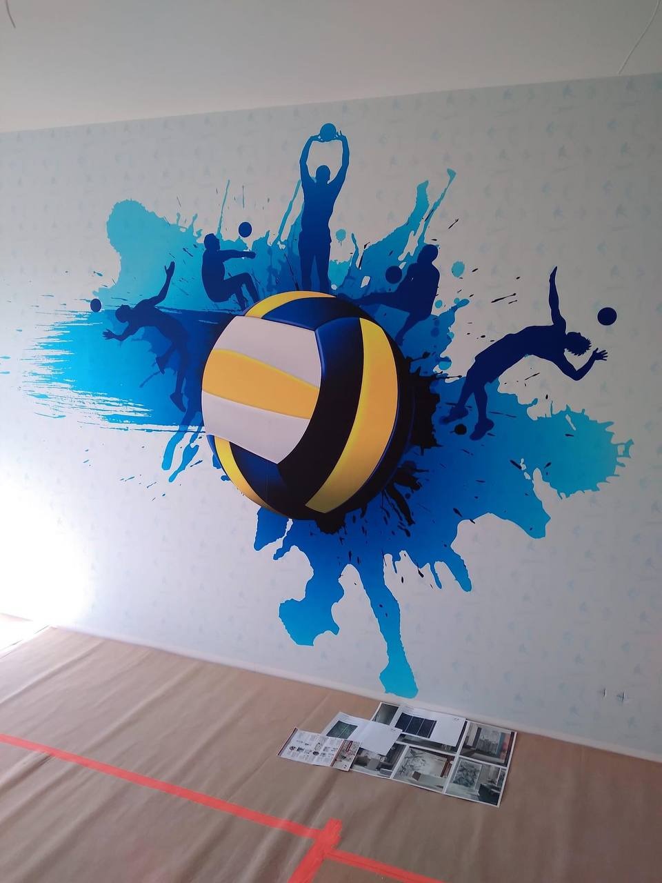 Ściana w Pruszkowie ozdobiona malunkiem przedstawiającym piłkę do siatkówki i sylwetki siatkarzy na tle niebieskich plam, zabezpieczona podłoga z widocznymi materiałami informacyjnymi.