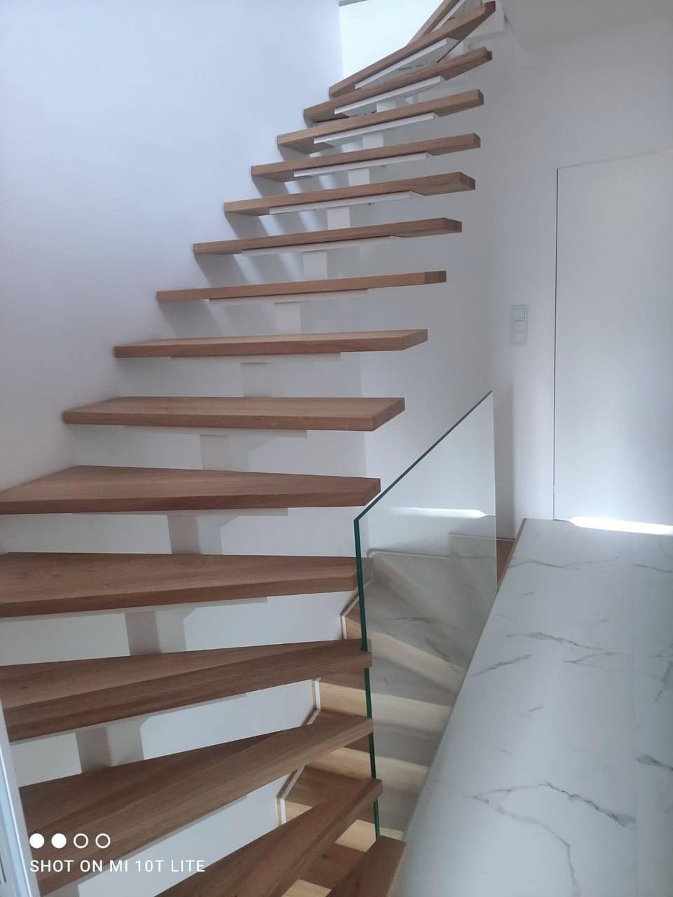 Nowoczesne drewniane schody samonośne z przezroczystą szklaną balustradą, zamontowane przy białej ścianie. Widoczne marmurowe wykończenie podłogi i białe podstopnice.
