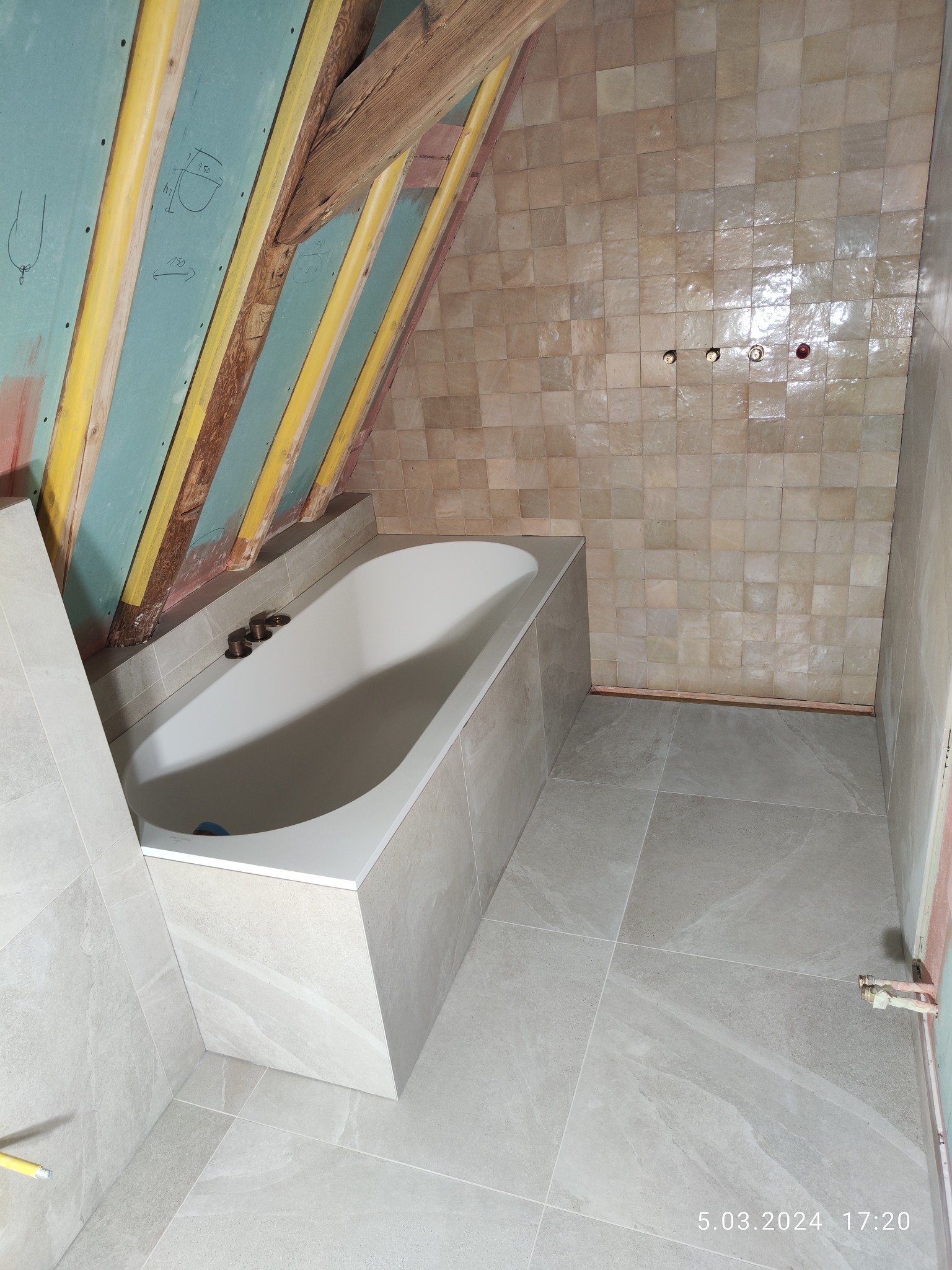 Łazienka na poddaszu w trakcie remontu: biała wanna obudowana płytkami, beżowe płytki na ścianach, drewniana konstrukcja dachu z zielonymi płytami kartonowo-gipsowymi z naniesionymi odręcznymi...