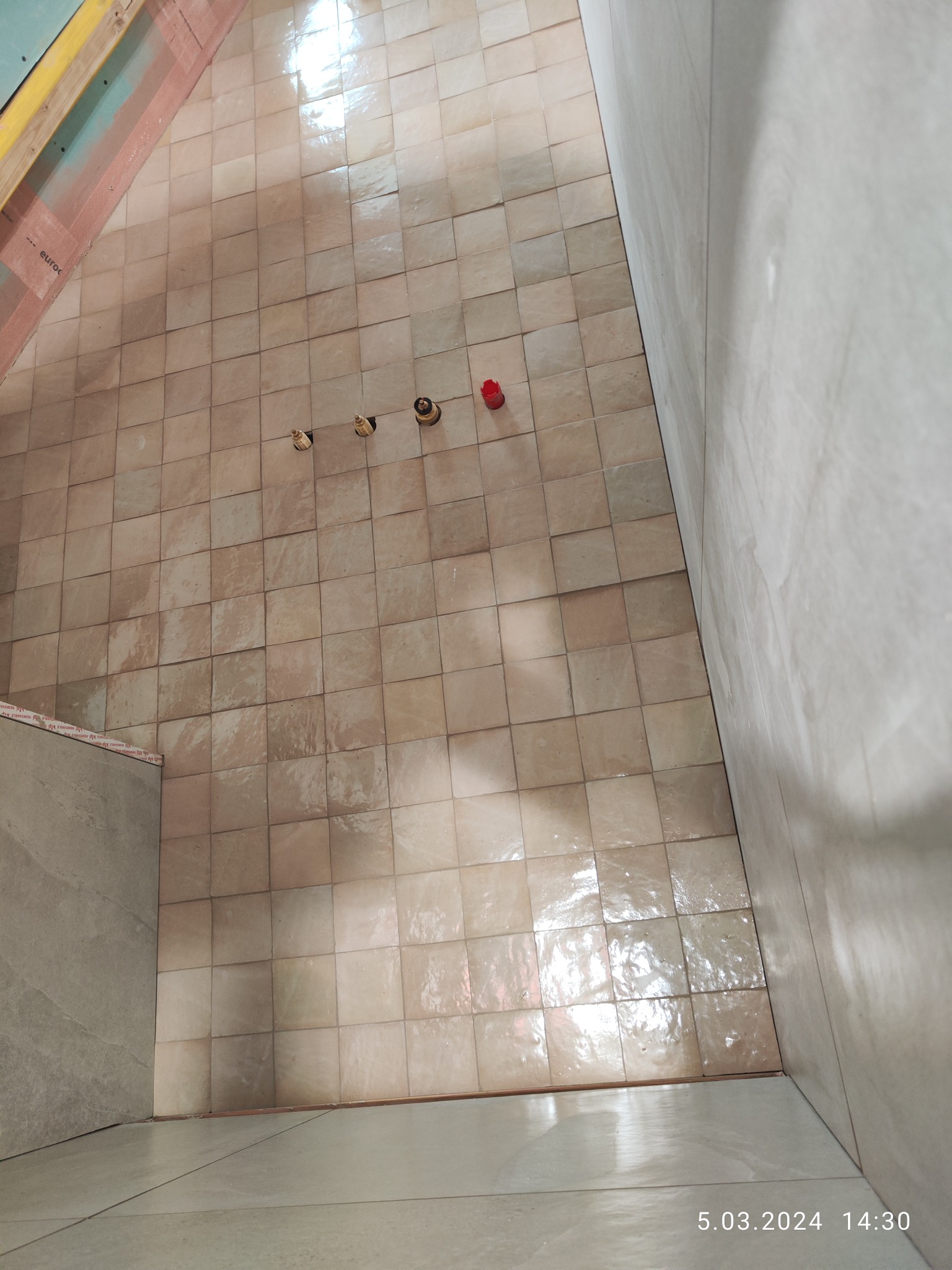 Podłoga łazienki wyłożona kwadratowymi, beżowymi płytkami ceramicznymi; widoczne trzy wystające rury instalacji wodnej i czerwona zaślepka, ściany w trakcie prac wykończeniowych.