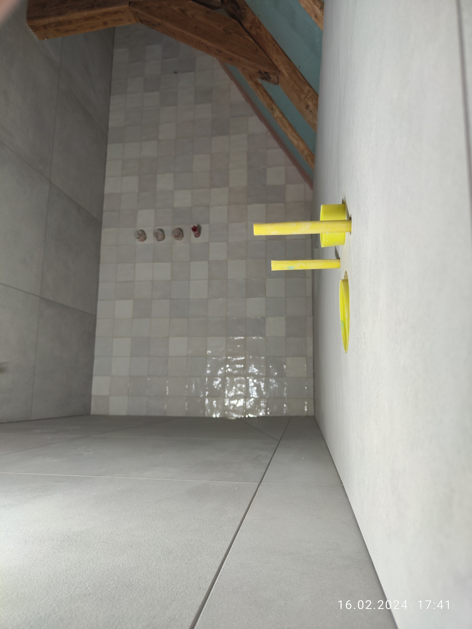 Świeżo położone, jasnoszare płytki podłogowe i ścienne w łazience, z widocznymi żółtymi rurami instalacji wodnej wystającymi ze ściany, surowy drewniany strop w tle.