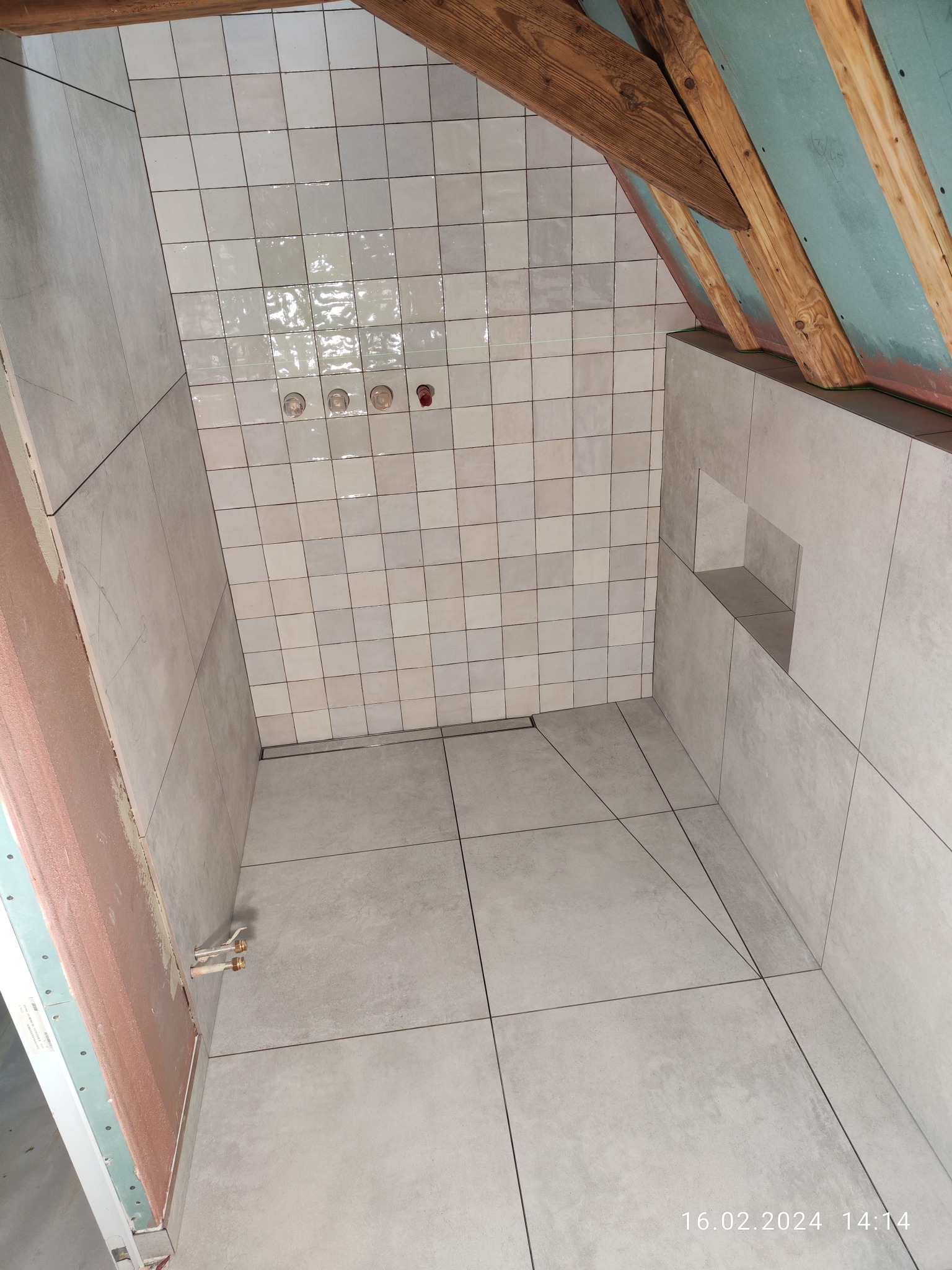 Wnętrze łazienki w trakcie remontu na poddaszu, widoczne szare płytki na ścianach i podłodze, surowe drewniane belki stropowe, instalacja wodna bez armatury i wnęka w ścianie.