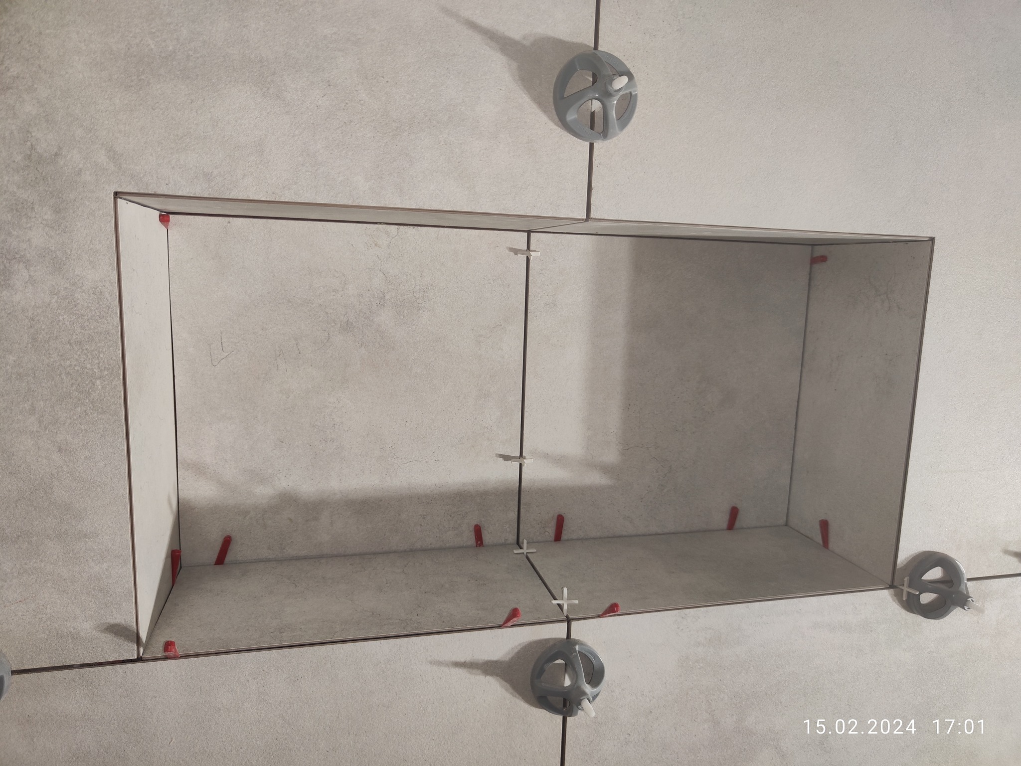 Wnęka wyłożona jasnymi płytkami ceramicznymi w trakcie prac, widoczne krzyżyki dystansowe i system poziomowania płytek, data i godzina w prawym dolnym rogu.