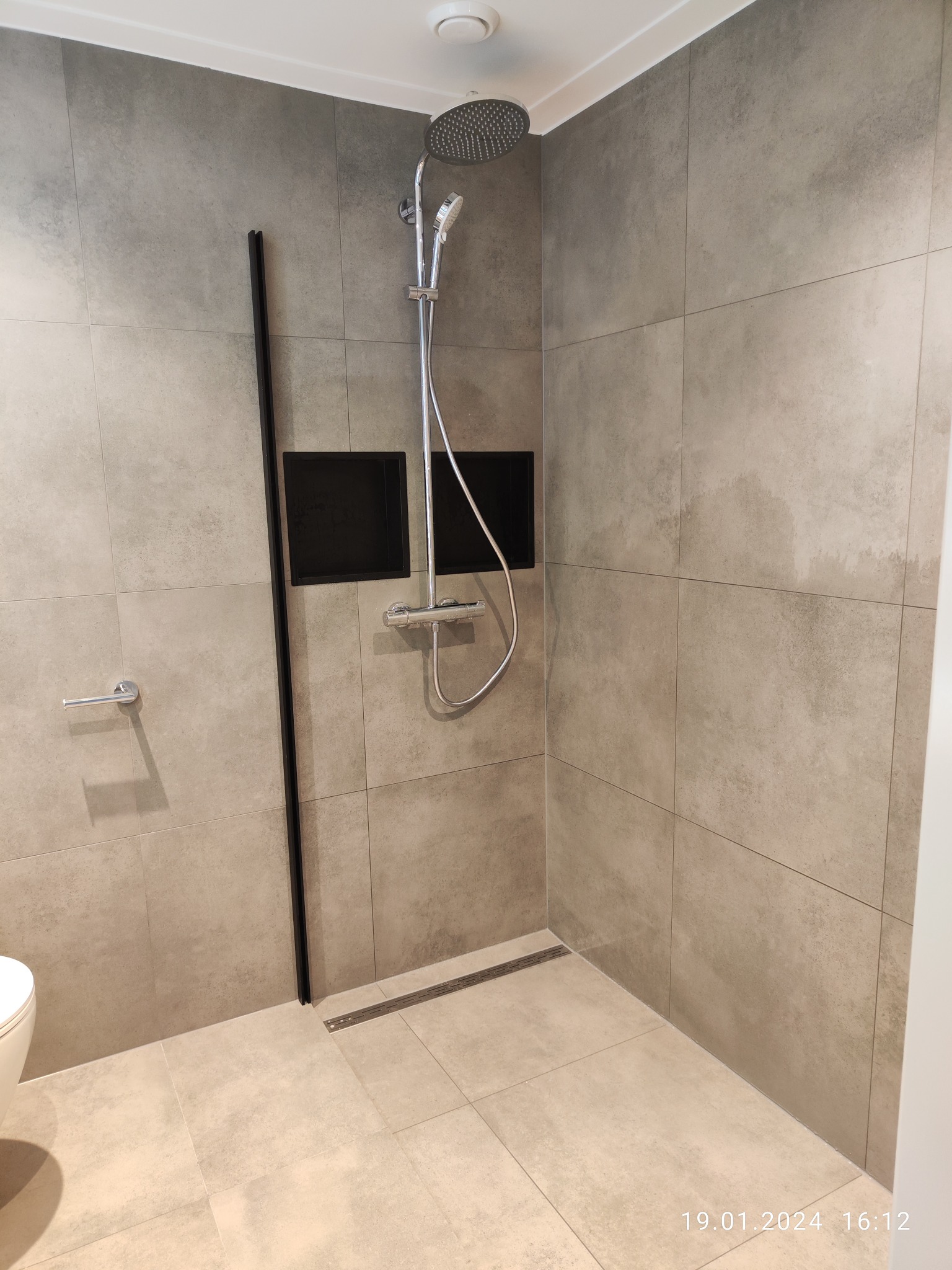 Wykończona kabina prysznicowa walk-in z odpływem liniowym, szare płytki wielkoformatowe na ścianach i podłodze, czarna rama szklanej ścianki, wnęki w ścianie z czarnym tłem, bateria termostatyczna.