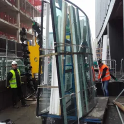 Montaż zakrzywionej szyby na placu budowy z użyciem przyssawki, pracownicy w kamizelkach ochronnych, widoczne rusztowania i budynek w tle, mokra nawierzchnia.