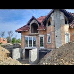 Bartosz Doszko... - Budynek w trakcie budowy z zamontowanymi białymi oknami, widoczne fundamenty i ceglana elewacja, teren wokół rozkopany.