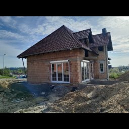 Bartosz Doszko... - Dom w trakcie budowy z zamontowanymi białymi oknami i drzwiami balkonowymi, widoczne cegły i czerwona dachówka, teren wokół domu w trakcie prac ziemnych.