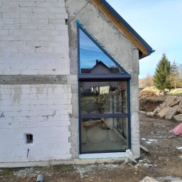 Bartosz Doszko... - Nietypowe okno o trójkątnym kształcie w surowym stanie budynku, z częściowo opuszczoną roletą zewnętrzną, odbijające krajobraz.