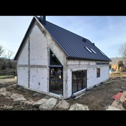 Bartosz Doszko... - Surowy stan budynku z oknami w czarnych ramach i roletą zewnętrzną częściowo opuszczoną na trójkątnym oknie, widoczne rusztowania wewnątrz, czarny dach i gruz przed budynkiem.