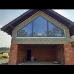 Surowy stan budynku z zamontowanym trójkątnym oknem z szarymi ramami, odbijającym drzewa i niebo; widoczny czerwony mur z cegieł i betonowy nadproże.