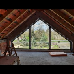 Bartosz Doszko... - Trójkątne okno z widokiem na zewnątrz, zamontowane w surowym stanie poddasza z drewnianą konstrukcją dachu i widocznym rusztowaniem.